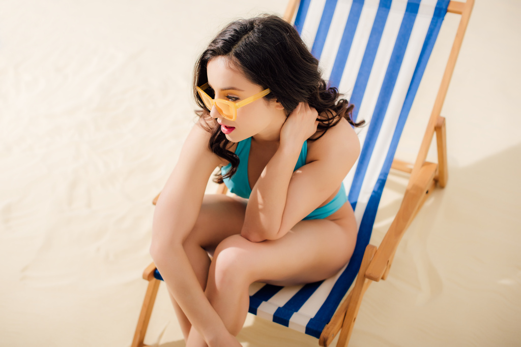 красивая девушка в бикини и солнцезащитных очках сидит на шезлонге на пляже
 - Фото, изображение