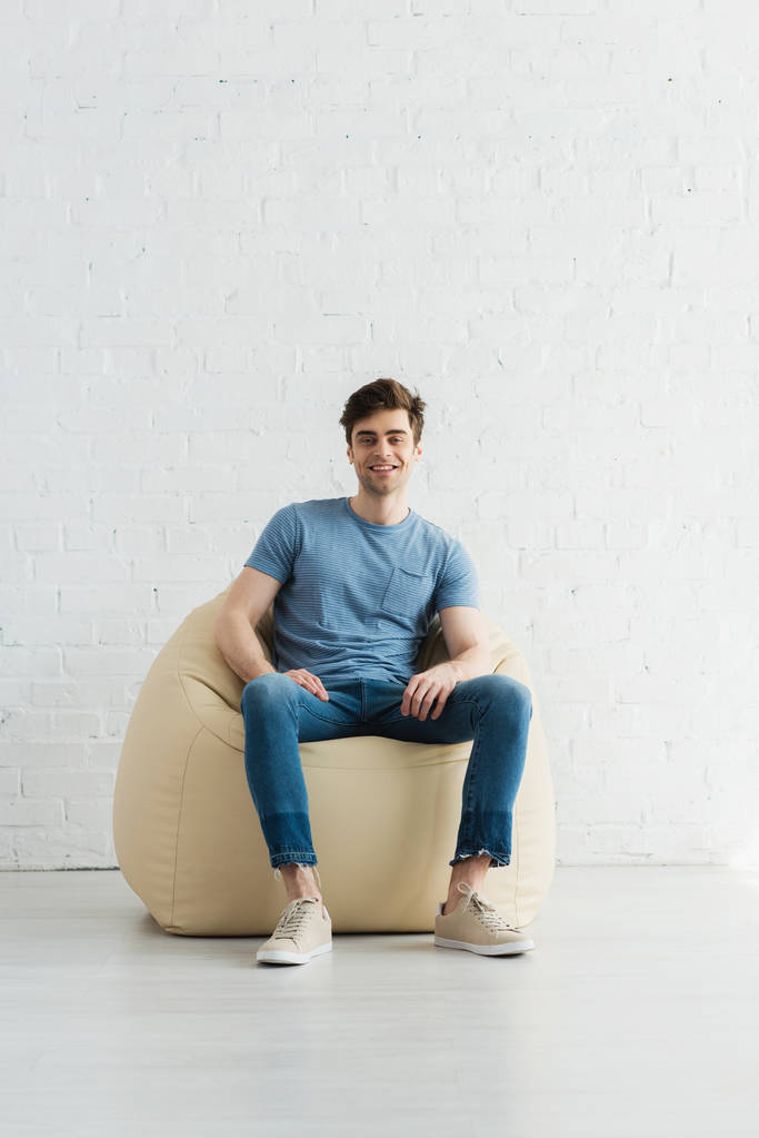 evde bej fasulye torbası sandalyede oturan mutlu ve yakışıklı adam  - Fotoğraf, Görsel