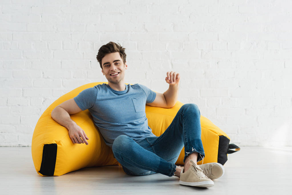 szczęśliwy i przystojny człowiek siedzi z skrzyżowanymi nogami w pobliżu żółty fasola torba krzesło w domu  - Zdjęcie, obraz