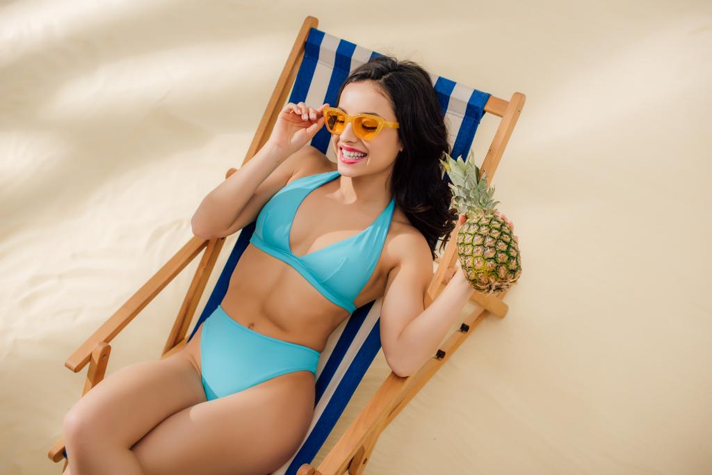 bela menina feliz em óculos de sol e biquíni segurando abacaxi e relaxante na cadeira deck na praia
 - Foto, Imagem