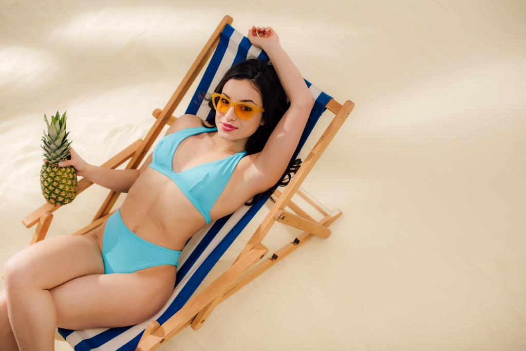 όμορφο κορίτσι σε γυαλιά ηλίου και μπικίνι κοιτάζοντας την κάμερα και κρατώντας ανανά, ενώ χαλάρωσηστο κατάστρωμα καρέκλα στην παραλία - Φωτογραφία, εικόνα