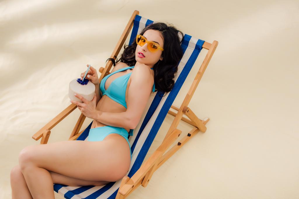 beautiful girl in bikini and sunglasses with coconut cocktail lying on deck chair and looking at camera on beach - Photo, Image