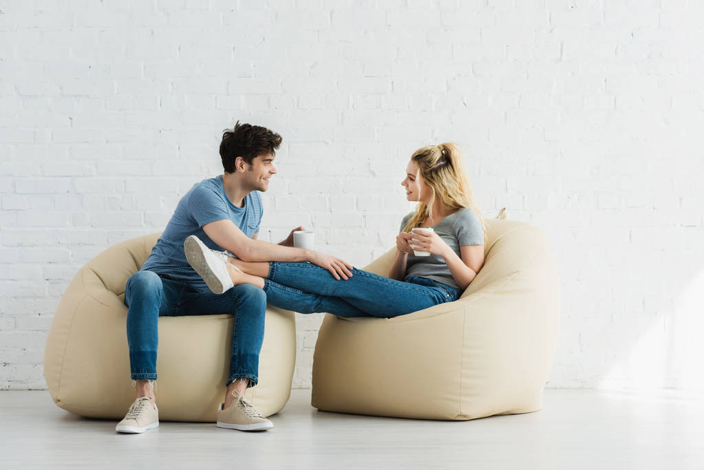 cheerful woman and happy man sitting on bean bag chairs and holding cups  - Photo, Image