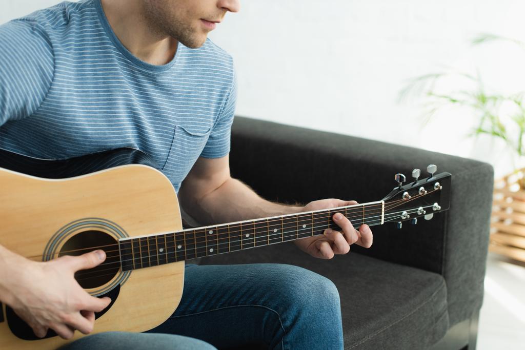 обрезанный вид музыканта, сидящего на диване и играющего на акустической гитаре дома
  - Фото, изображение
