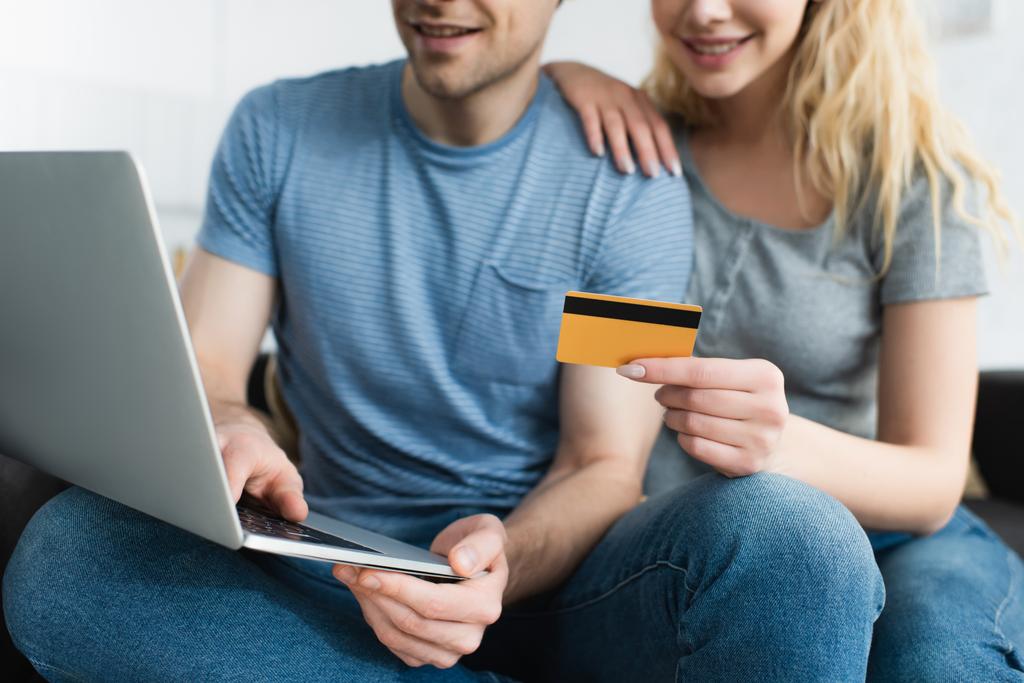 περικομμένη θέα της ξανθιά γυναίκα που κατέχουν πιστωτική κάρτα κοντά χαρούμενο άντρα χρησιμοποιώντας φορητό υπολογιστή - Φωτογραφία, εικόνα