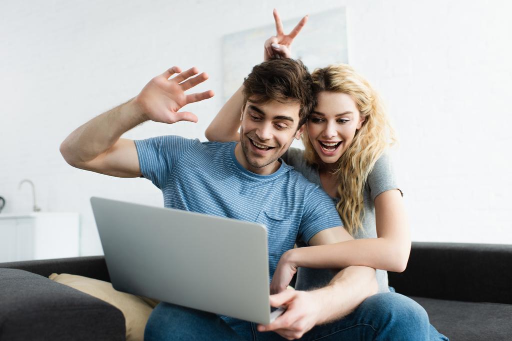 χαρούμενος άνθρωπος κουνώντας το χέρι κοντά χαρούμενη γυναίκα δείχνοντας το σήμα της ειρήνης, ενώ έχοντας βίντεο κλήση στο laptop  - Φωτογραφία, εικόνα