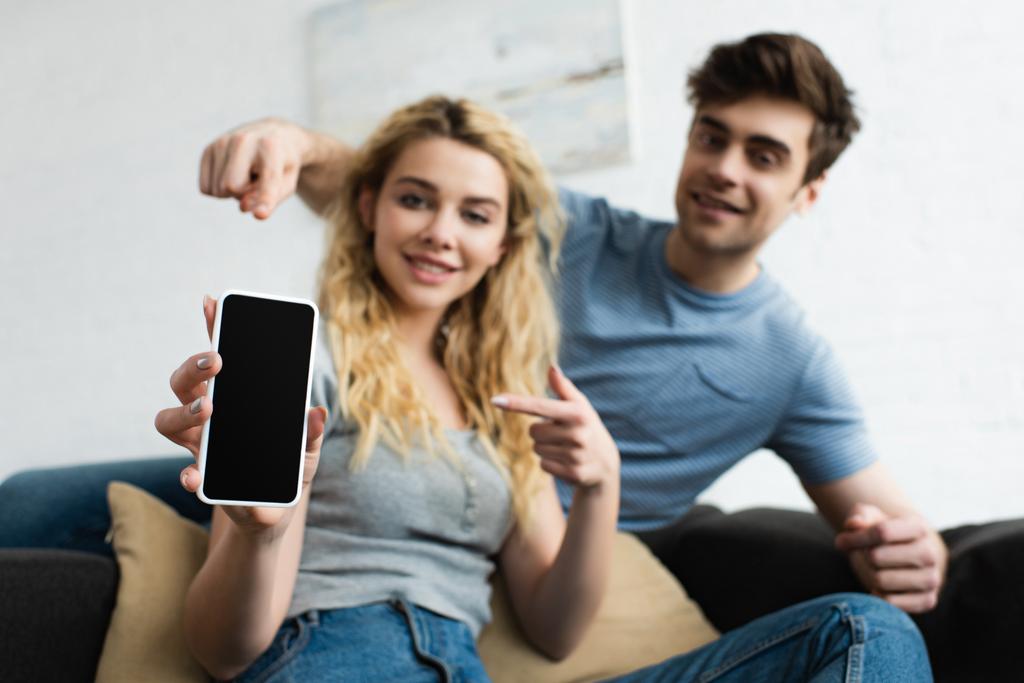 selective focus of cheerful man and blonde woman pointing with fingers at smartphone with blank screen - Photo, Image