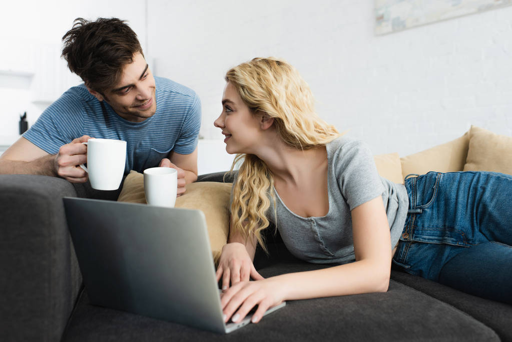 heureux et beau homme tenant tasses près fille blonde en utilisant un ordinateur portable
  - Photo, image
