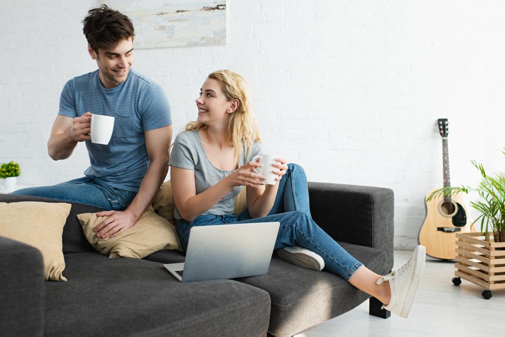happy man holding cup of coffee and sitting on sofa near cheerful blonde girl with laptop - Photo, Image