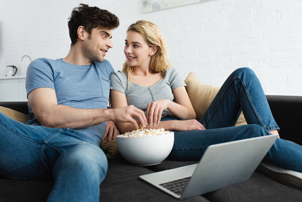 happy man and cheerful blonde woman taking popcorn from bowl near laptop  - Photo, Image