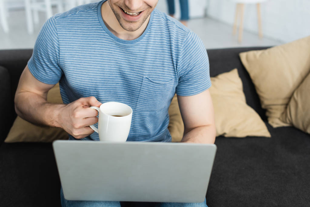 rajattu näkymä onnellinen mies käyttää kannettavaa tietokonetta pitäen cup juomalla
  - Valokuva, kuva