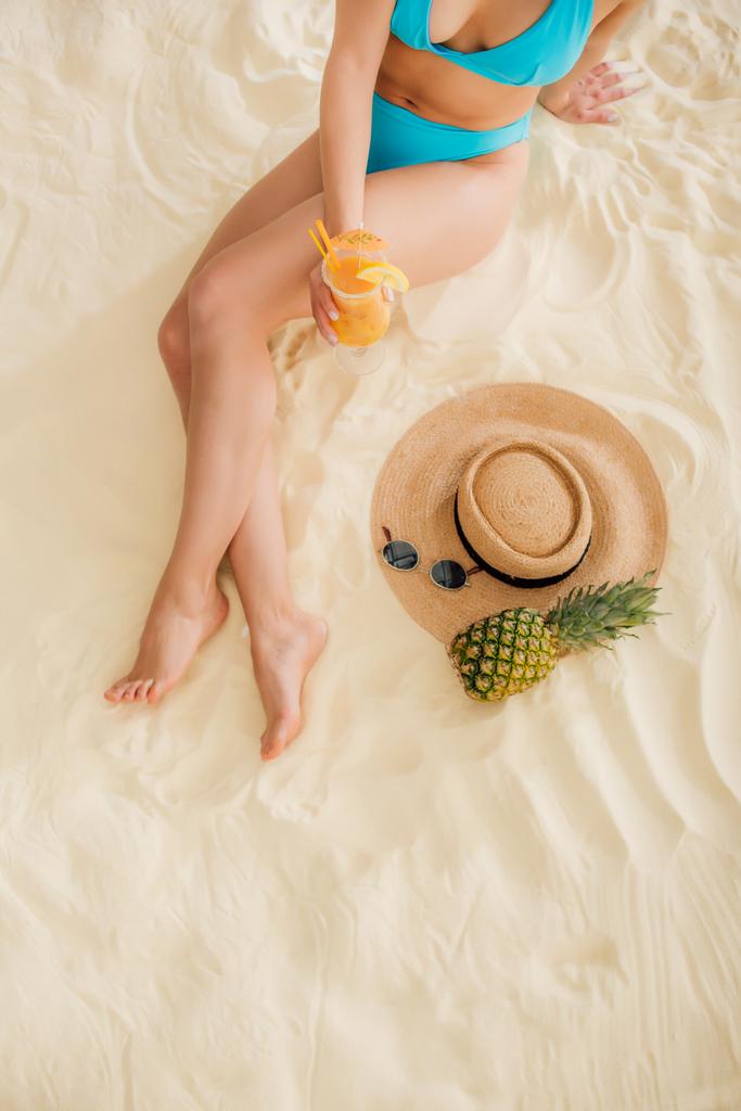 Top uitzicht van meisje in bikini met cocktail, stro hoed, ananas en zonnebril op strand - Foto, afbeelding