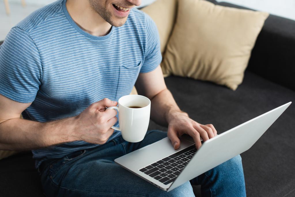 vista ritagliata dell'uomo allegro che utilizza il computer portatile mentre tiene la tazza con la bevanda
  - Foto, immagini