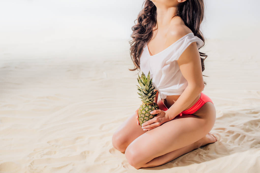 vista recortada de la niña sentada y posando con piña en la playa de arena
 - Foto, Imagen