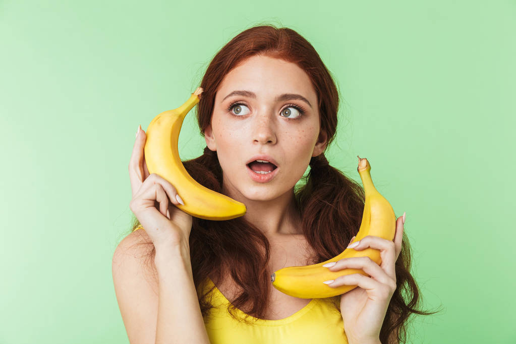 バナナの果物と緑の壁の背景の上に孤立した若い赤毛の女の子のポーズ. - 写真・画像
