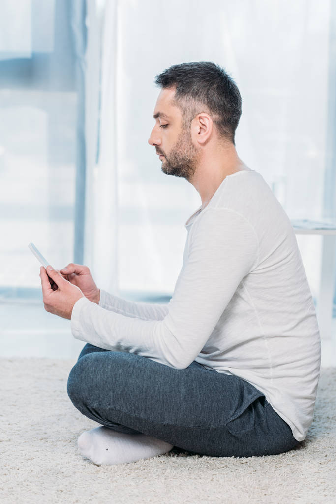 boční pohled na pohledného muže v příležitostném oblečení, které sedí na koberci a používají smartphone doma - Fotografie, Obrázek