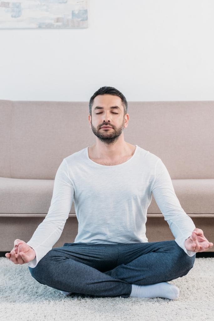 ロータスポーズでカーペットの上に座って、自宅で瞑想する目を閉じたハンサムな男 - 写真・画像