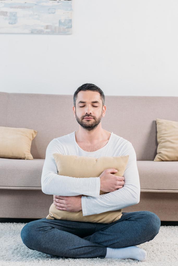 bonito homem com olhos fechados sentado no tapete e abraçando travesseiro em casa na sala de estar
 - Foto, Imagem
