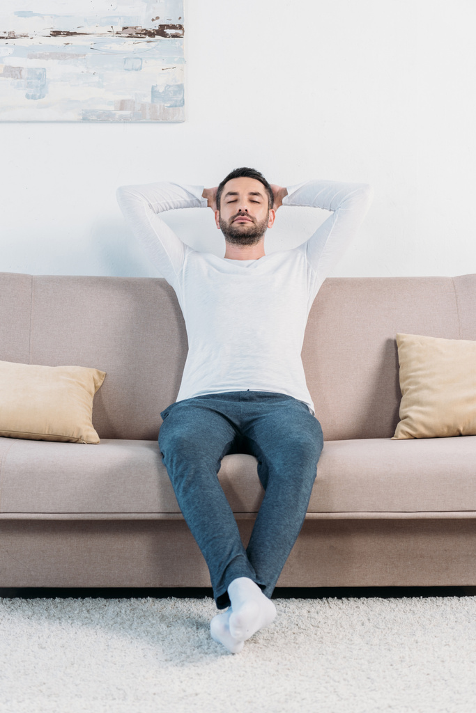 bonito barbudo homem com olhos fechados e mãos atrás das costas descansando no sofá em casa
 - Foto, Imagem