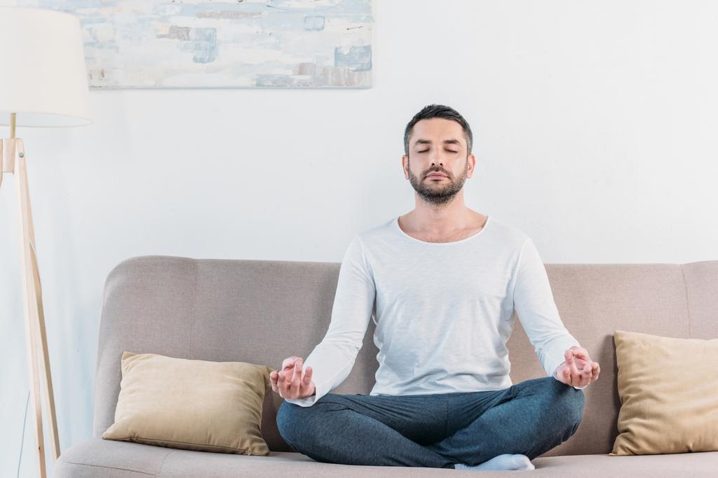 schöner Mann mit geschlossenen Augen sitzt auf Couch in Lotus-Pose und meditiert zu Hause - Foto, Bild
