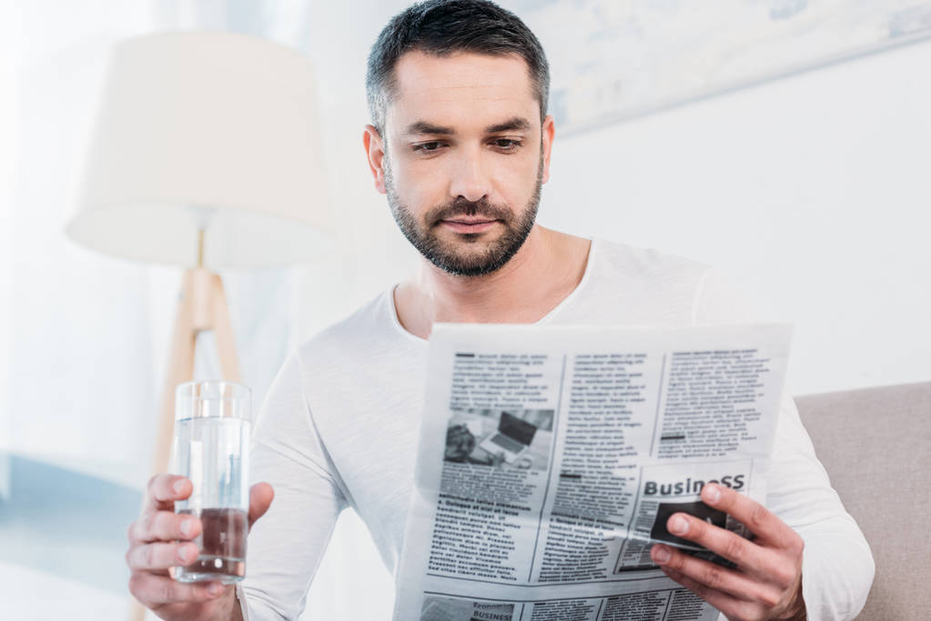 handsome bearded man sitting with glass of water and reading newspaper at home - Photo, Image