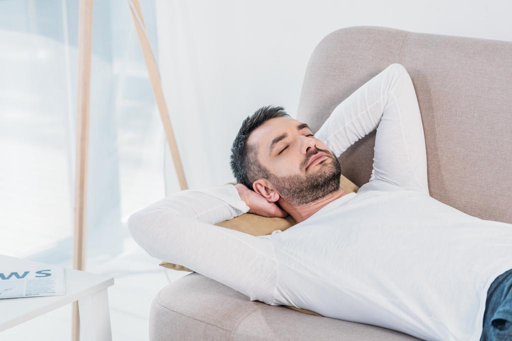 красивый бородатый мужчина с закрытыми глазами и руками за спиной спит на диване дома
 - Фото, изображение