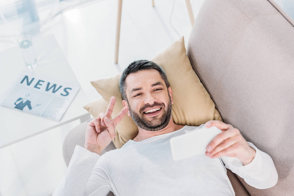 komea hymyilevä mies makaa sohvalla ja ottaa selfie älypuhelimeen kotona
 - Valokuva, kuva
