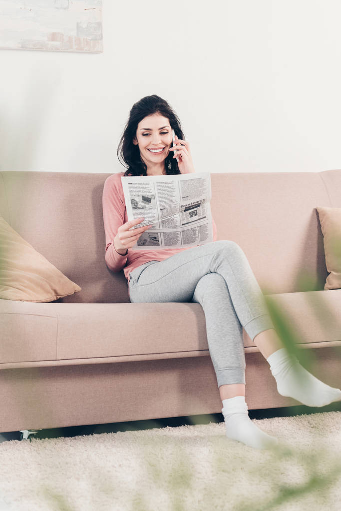 selektivní zaměření krásné usměvavé ženy sedící na pohovce, čtení novin a používání smartphone doma - Fotografie, Obrázek