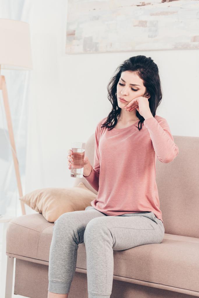 sad woman sitting on couch with glass of water and touching face at home - Photo, Image