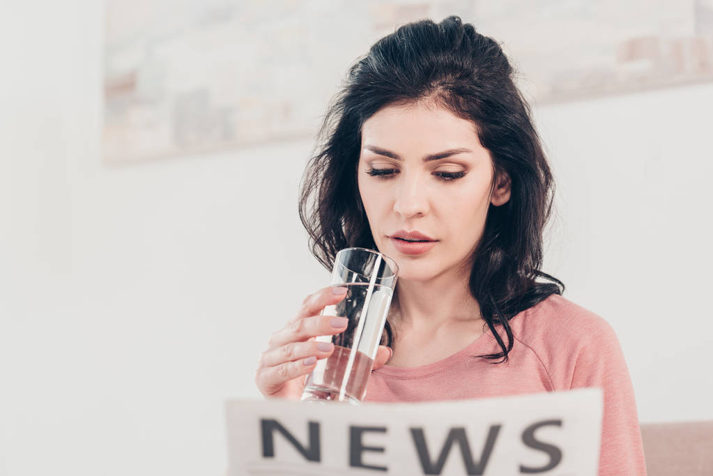 beautiful woman holding glass of water and reading newspaper at home - Photo, Image