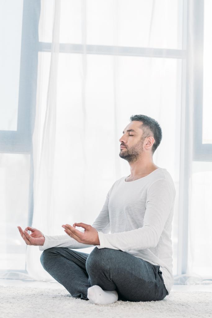 schöner Mann mit geschlossenen Augen sitzt auf Teppich in Lotus-Pose und meditiert zu Hause - Foto, Bild