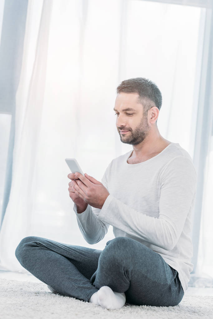 όμορφος άντρας σε casual ρούχα κάθεται στο χαλί και χρησιμοποιώντας το smartphone στο σπίτι με το φως του ήλιου - Φωτογραφία, εικόνα