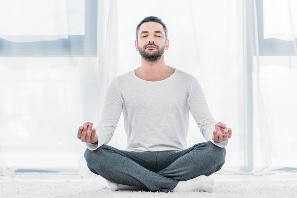 dobrze wyglądający człowiek z zamkniętymi oczami siedzi na dywanie w Lotus Pose i medytuje w domu - Zdjęcie, obraz