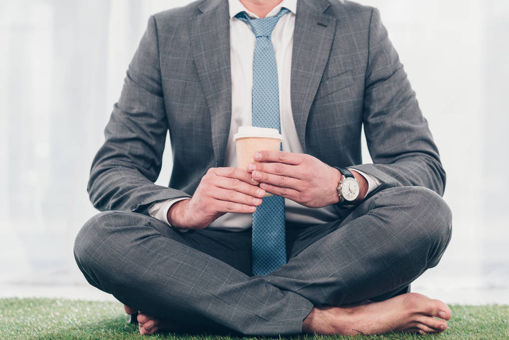 cropped view of businessman in suit with coffee to go sitting on grass mat - Photo, Image