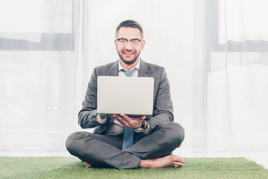 handsome smiling businessman in suit sitting on grass mat and using laptop - Photo, Image