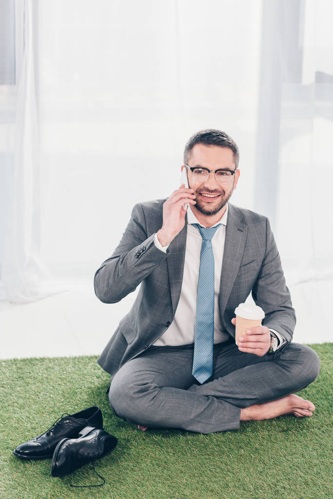 красивий усміхнений бізнесмен в костюмі сидить на траві мат, розмовляє на смартфоні і тримає каву, щоб піти
 - Фото, зображення