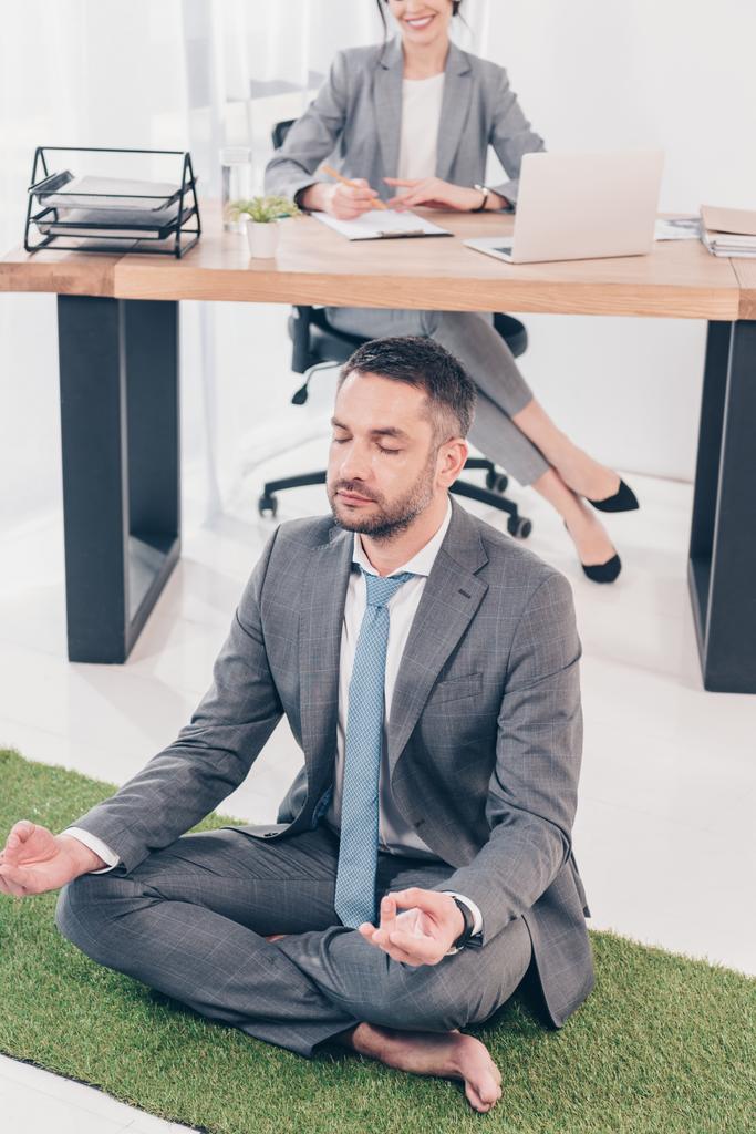 homme d'affaires méditant dans Lotus Poser sur tapis d'herbe tandis que la femme d'affaires assis à la table dans le bureau
 - Photo, image
