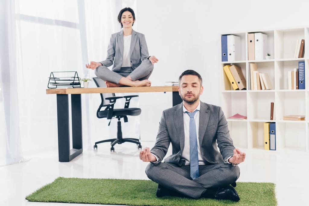 businessman meditating on grass mat while businesswoman sitting on table in Lotus Pose in office - Photo, Image