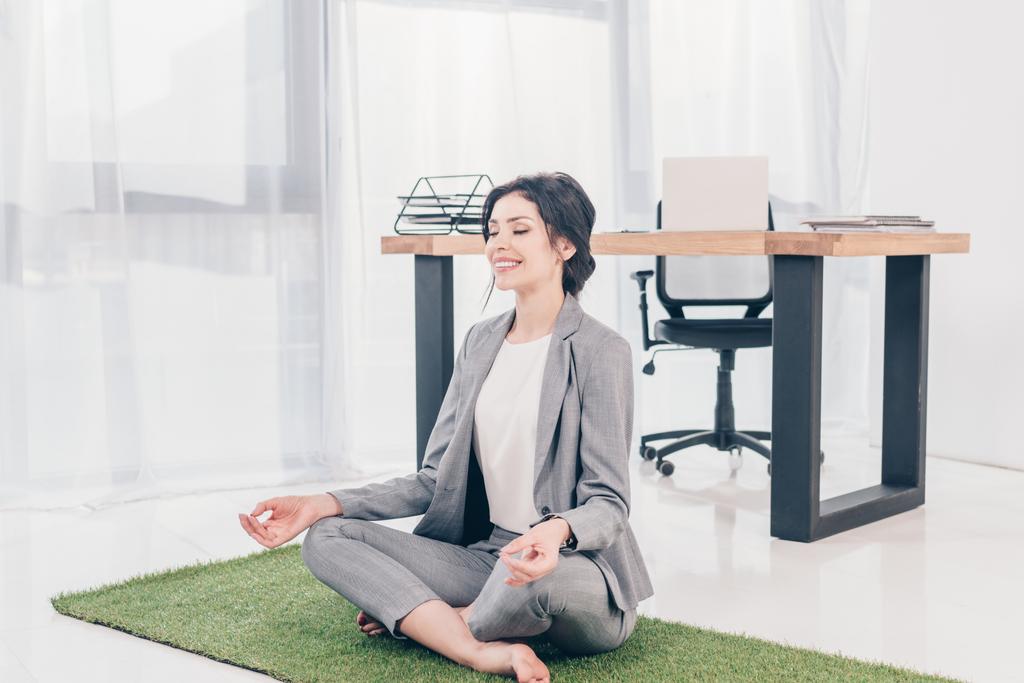 piękny szczęśliwy bizneswoman w garnitur siedzący na trawie Mata w Lotus Pose i medytowanie w biurze - Zdjęcie, obraz