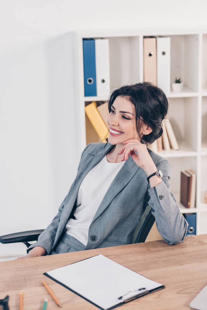 όμορφη χαμογελαστή επιχειρηματίας με κοστούμι κάθεται στο γραφείο στο γραφείο  - Φωτογραφία, εικόνα