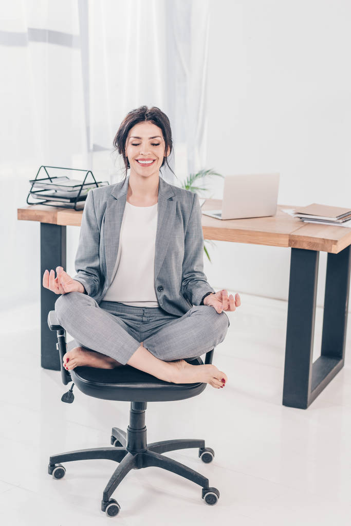 krásná usměvavá podnikatelka v obleku sedí na židli a medituje v Lotus Pose v úřadu - Fotografie, Obrázek
