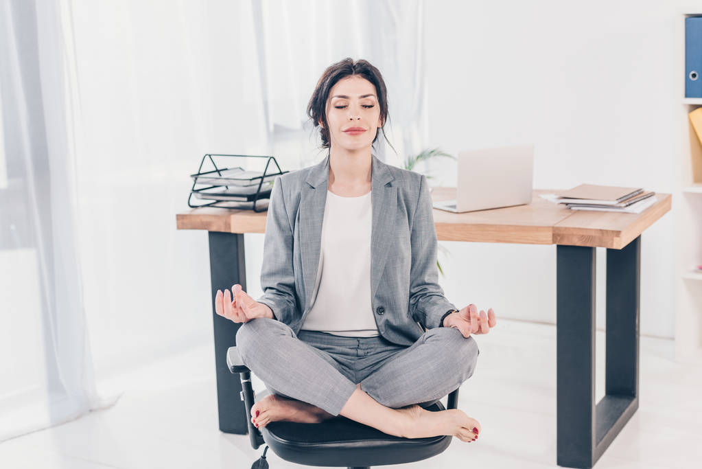 piękne businesswoman w garnitur siedzący na krześle i medytowanie w Lotus Pose w biurze - Zdjęcie, obraz