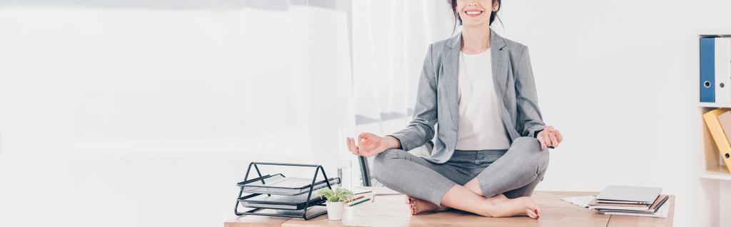 панорамный снимок деловой женщины в костюме, сидящей на столе и медитирующей в Lotus Pose в офисе
 - Фото, изображение