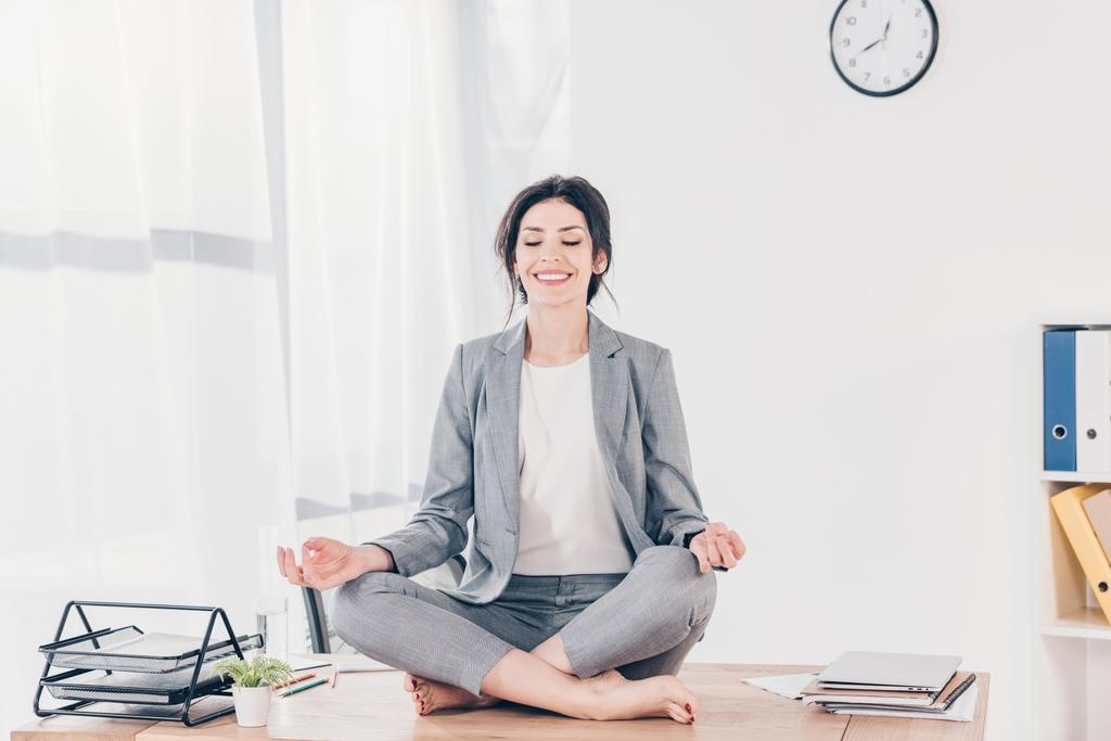 schöne lächelnde Geschäftsfrau im Anzug sitzt am Schreibtisch und meditiert in Lotus-Pose im Büro - Foto, Bild