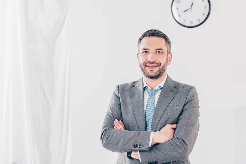 красивый улыбающийся бизнесмен в костюме со скрещенными руками смотрит на камеру в офисе с копировальным пространством
 - Фото, изображение