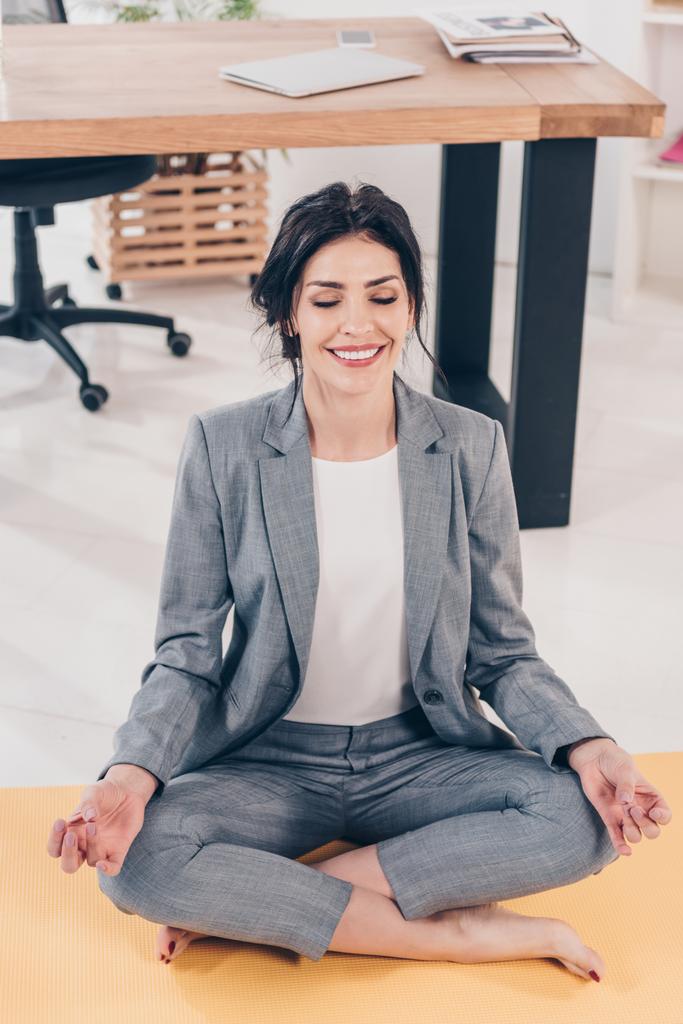 красивая улыбающаяся деловая женщина в костюме медитирует в Lotus Pose в офисе
 - Фото, изображение
