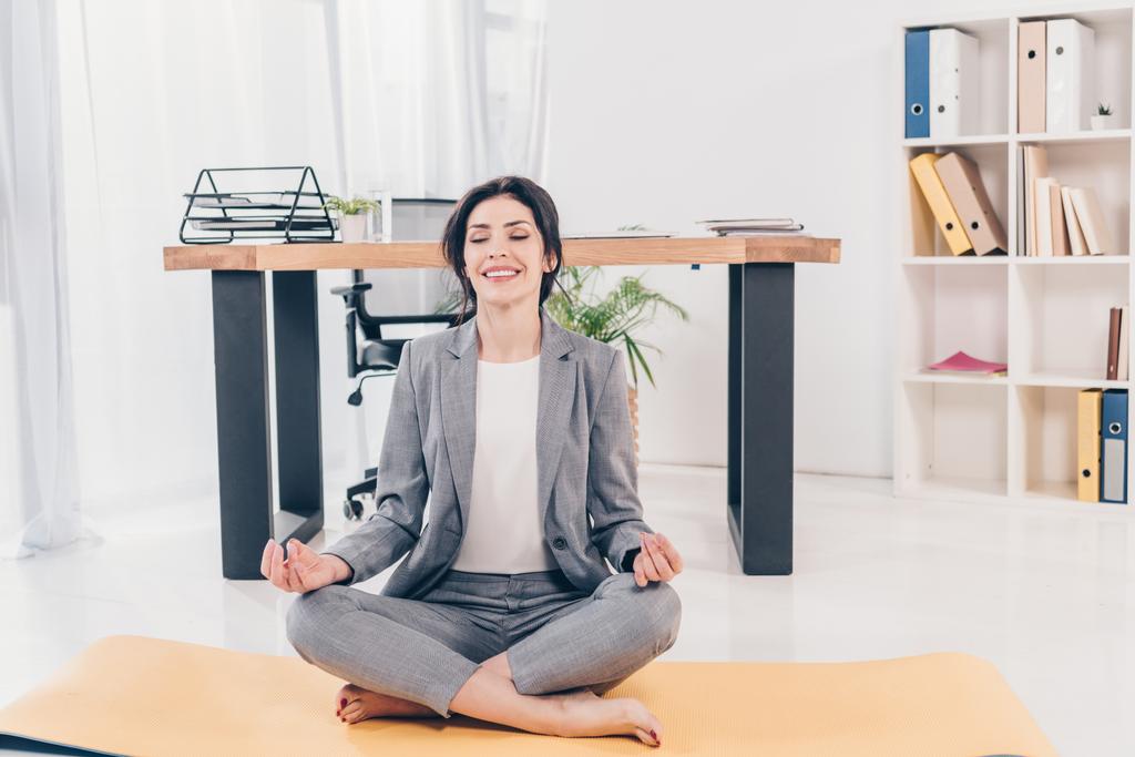 krásná usměvavá podnikatelka v obleku sedí na fitness podložce a medituje v kanceláři - Fotografie, Obrázek