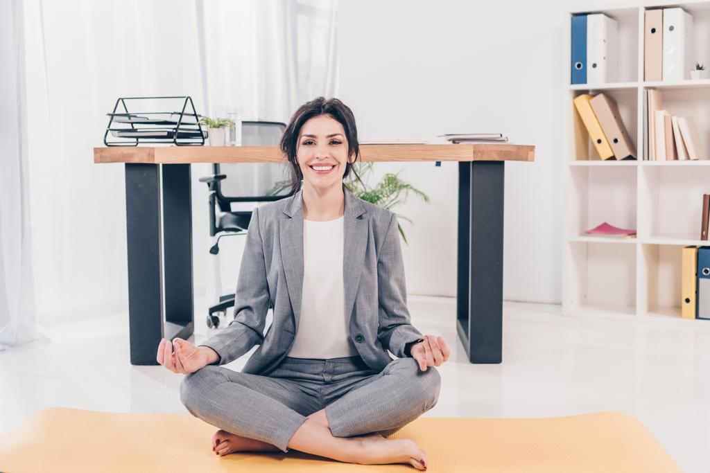 красивая улыбающаяся деловая женщина в костюме сидит на тренажерном коврике, смотрит в камеру и медитирует в офисе
 - Фото, изображение