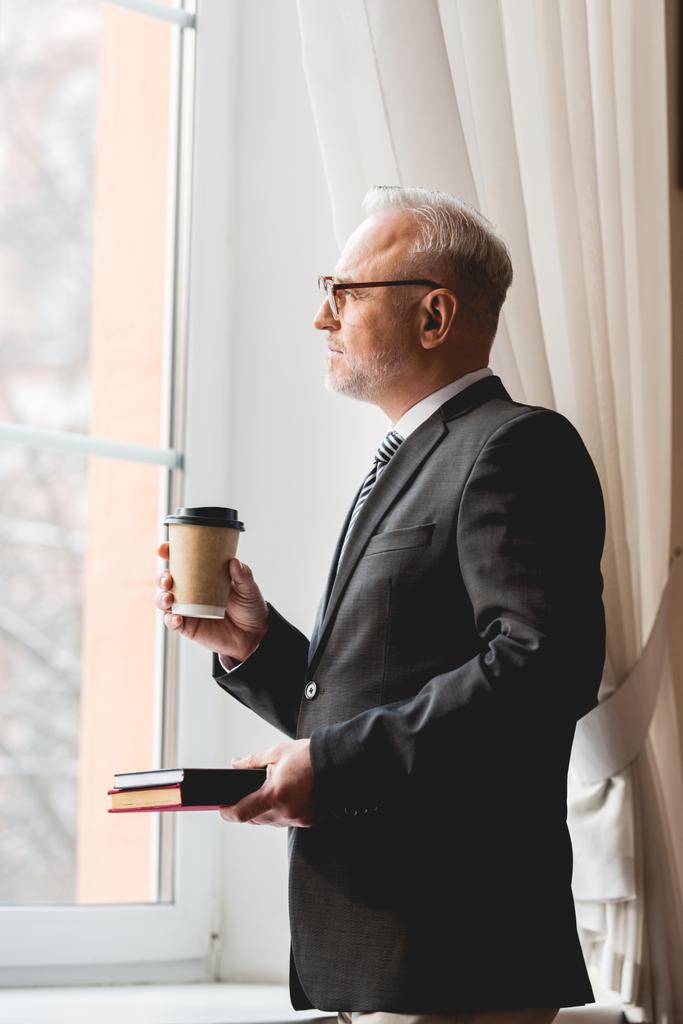 pensive mature teacher in glasses holding book and paper cup while looking at window  - Photo, Image
