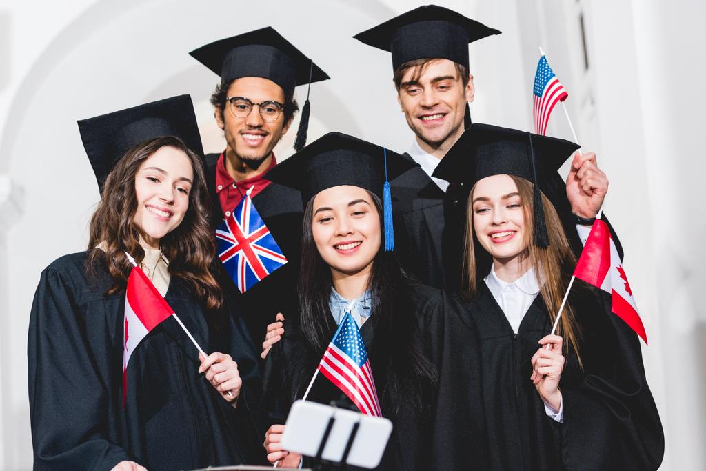 enfoque selectivo de los estudiantes alegres en vestidos de graduación con banderas de diferentes países y tomar selfie en el teléfono inteligente
  - Foto, Imagen